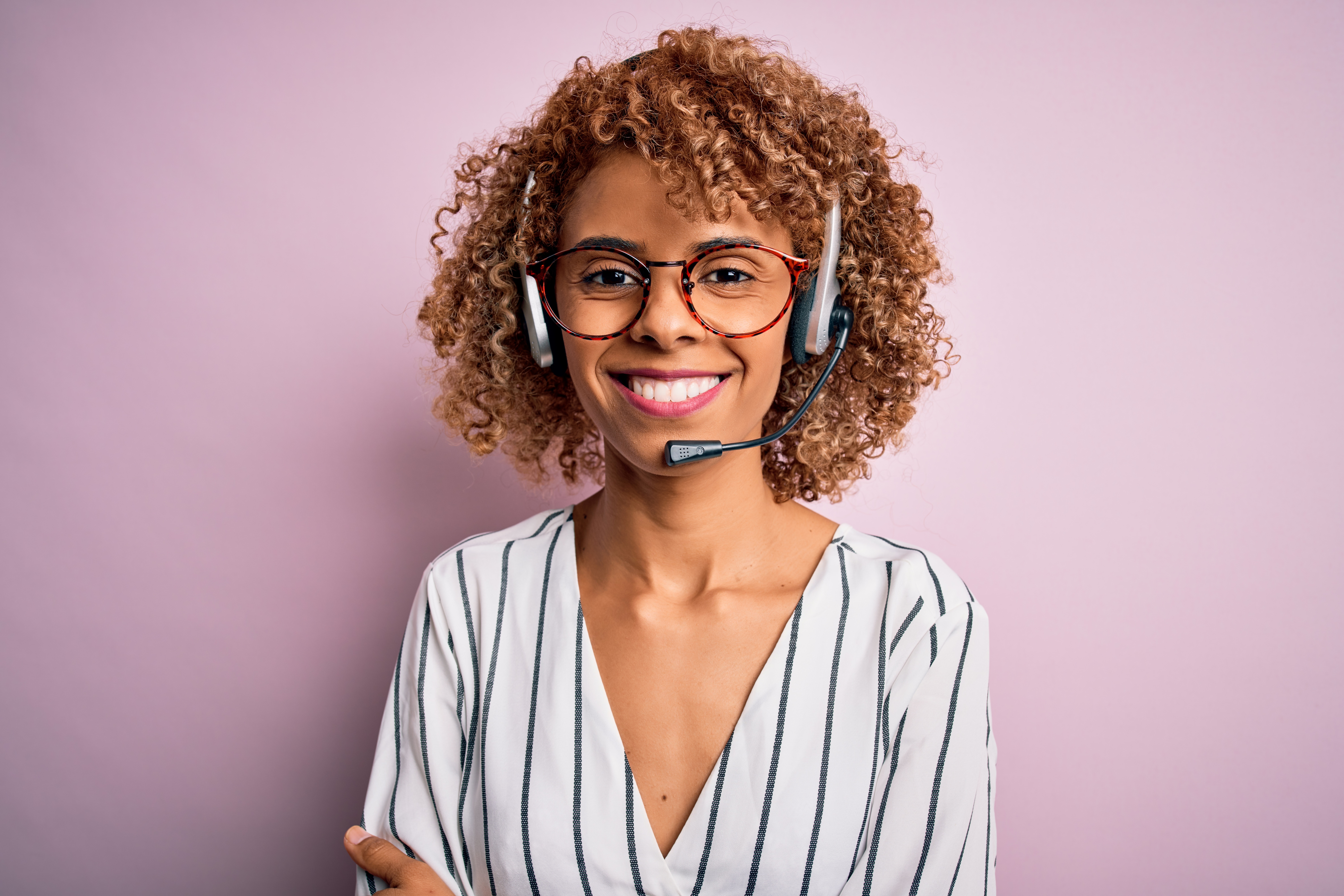African american curly call center agent woman working using headset over pink background happy face smiling with crossed arms looking at the camera. Positive person.